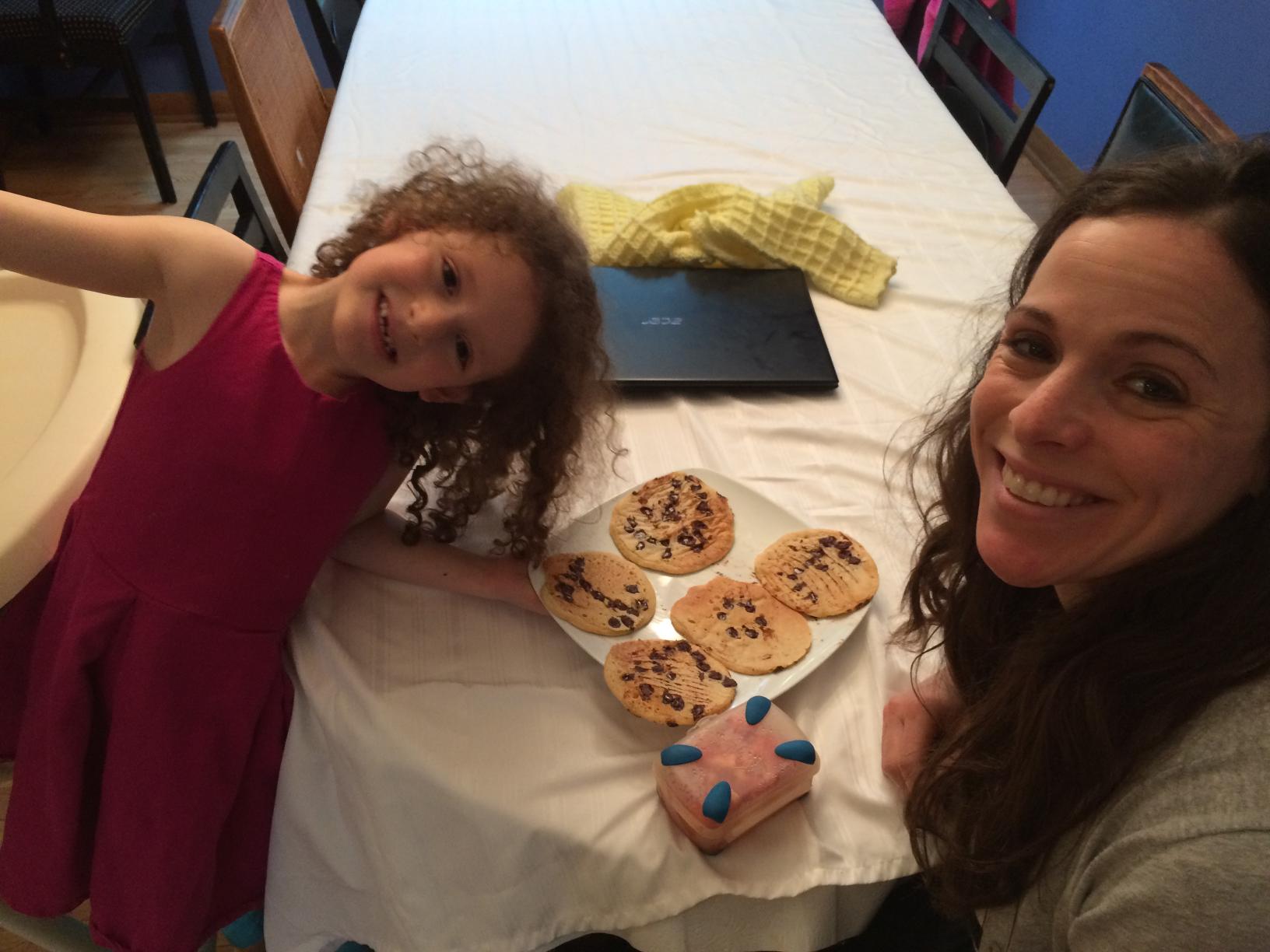 Smiling woman and young girl sit at a table with a plate of cookies and a pink frosted pastry in the foreground.