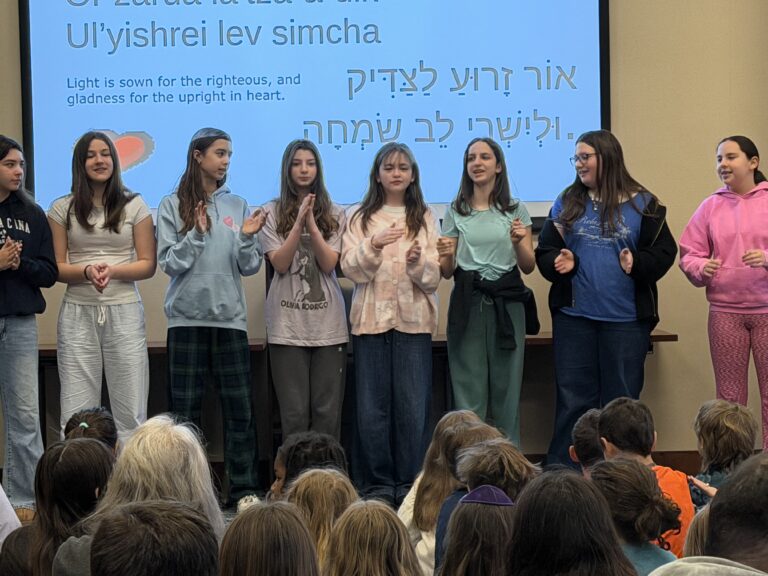 Group of young students standing on a stage, singing to a seated audience with a blue screen behind showing text in multiple languages.