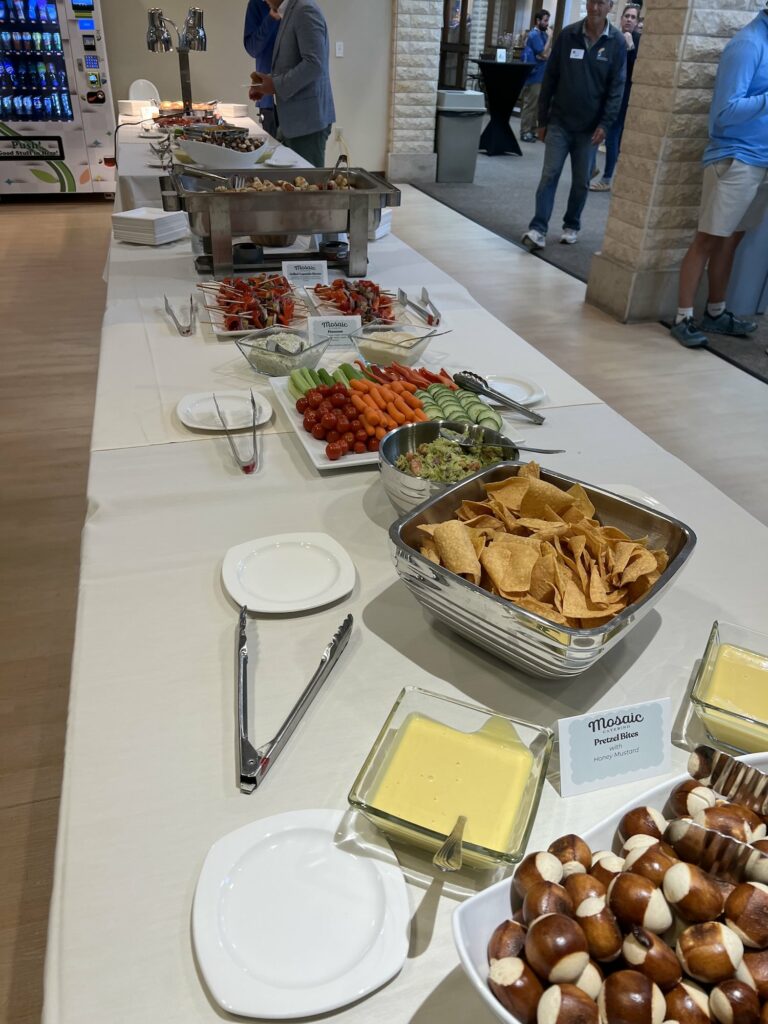 Buffet table with assorted snacks: cherry tomatoes, cucumbers, carrots, dips, skewers, and chips, in a conference setting behind a man in a blue jacket.