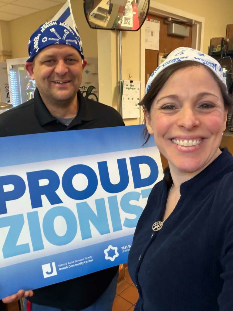 Smiling woman takes a selfie with a man holding a blue and white 'PROUD ZIONISTS' sign inside a community center. Both wear blue bandanas and casual clothing.