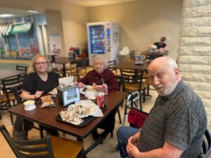 Man in foreground taking a selfie at a cafe table while two women sit nearby and snack; vending machine and seating area in the background.
