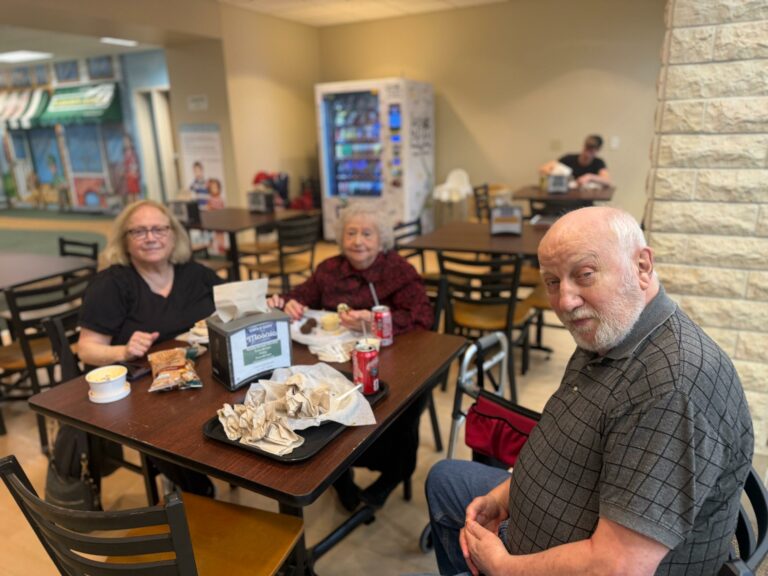Man in foreground taking a selfie at a cafe table while two women sit nearby and snack; vending machine and seating area in the background.