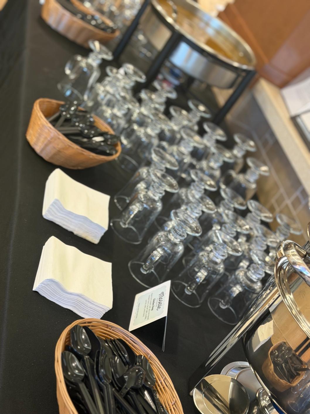 Buffet setup with many upside-down wine glasses arranged on a black tablecloth, plus baskets of spoons and napkins nearby.