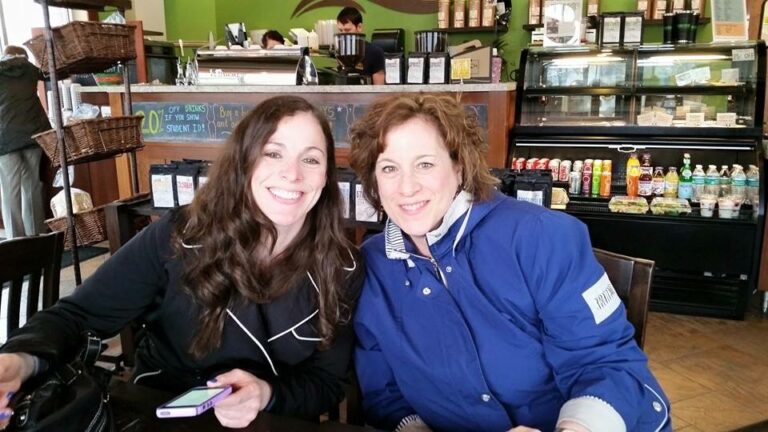 Two women sit close together in a cafe, smiling at the camera with drinks and a counter behind them.