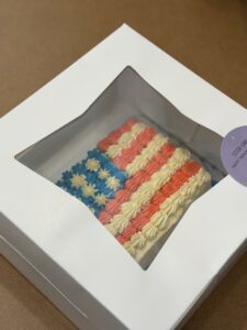 Boxed rainbow-frosted cake with pink, blue, and white swirls visible through a window in a white bakery box