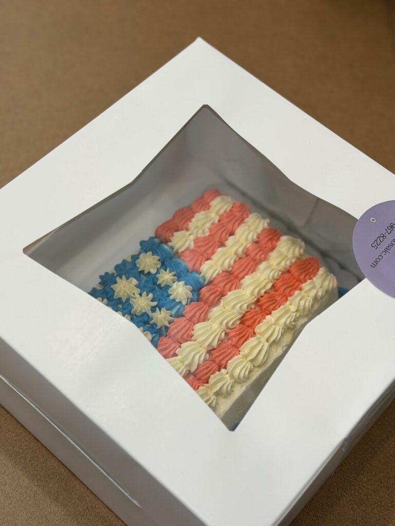 Boxed rainbow-frosted cake with pink, blue, and white swirls visible through a window in a white bakery box