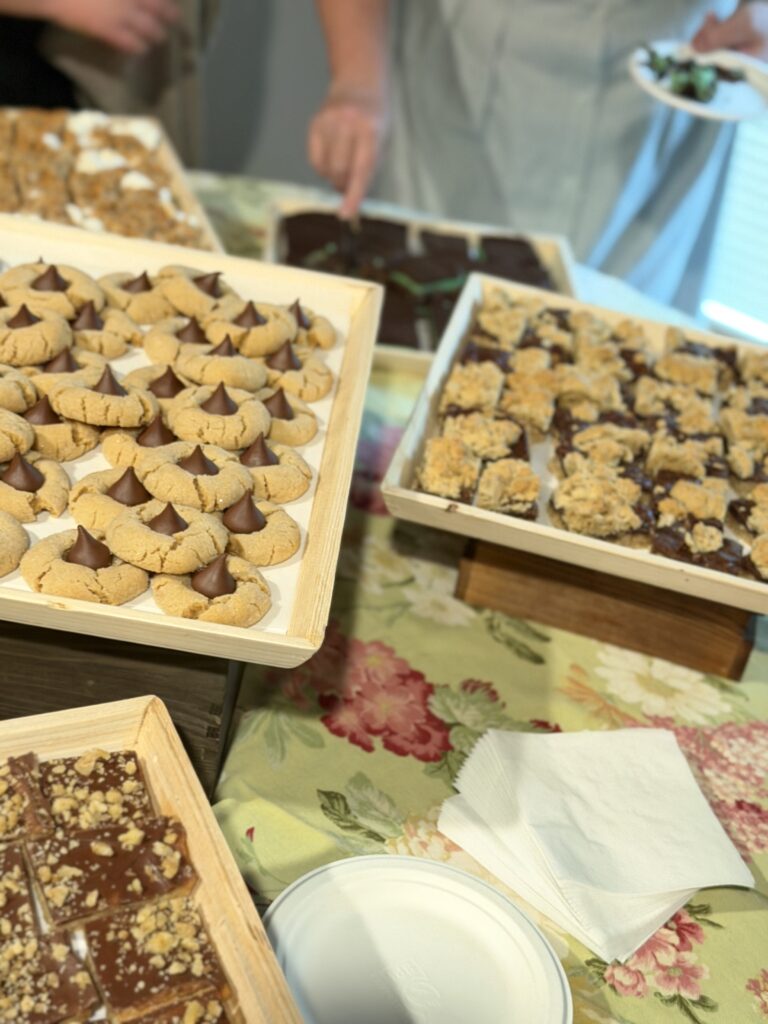 Tray of peanut butter blossom cookies with chocolate centers on a wooden platter at a dessert table, with other treats in the background.