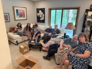 Family and friends lounging on a beige sectional in a living room, with open cardboard boxes on the floor nearby.