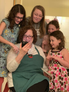 A smiling woman in a teal apron sits and shows a phone to a group of children gathered around her.