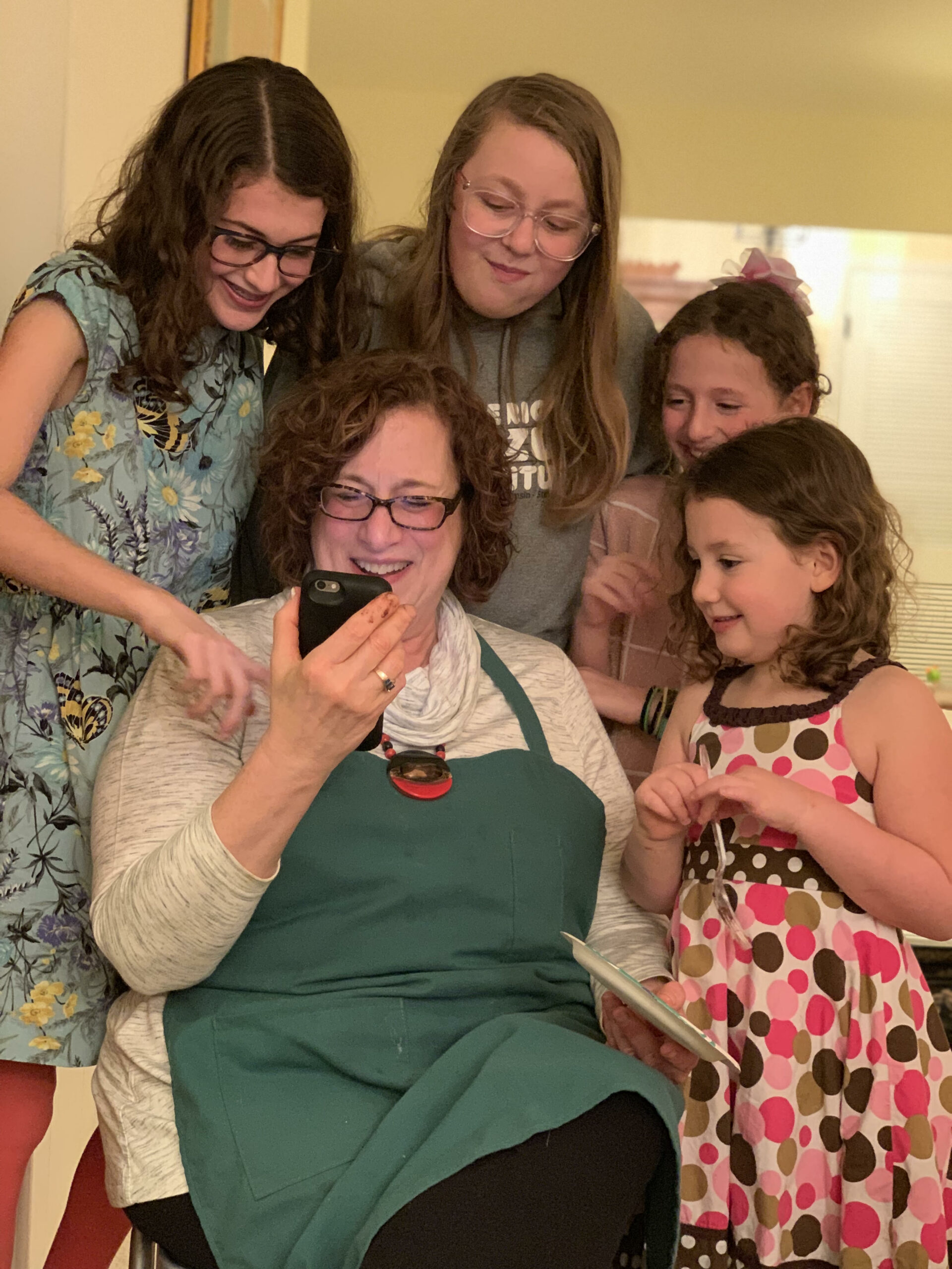 A smiling woman in a teal apron sits and shows a phone to a group of children gathered around her.