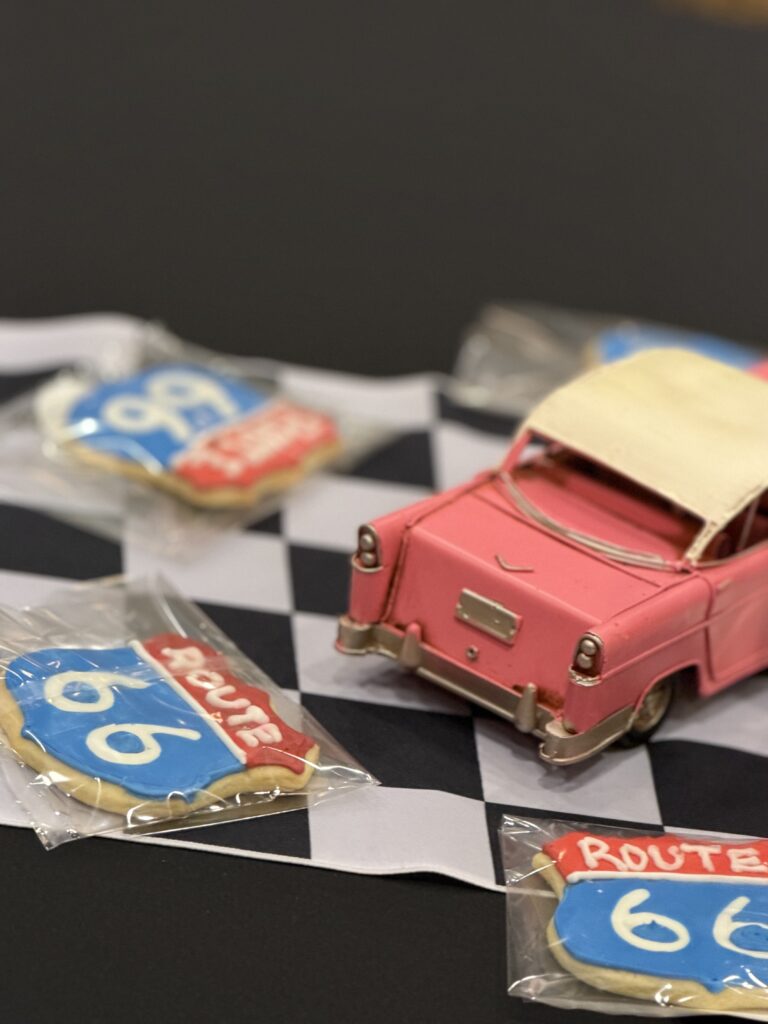 Pink toy car on a black-and-white checkered surface with packaged cookies in the foreground and blurred sweets nearby, creating a playful still life scene.