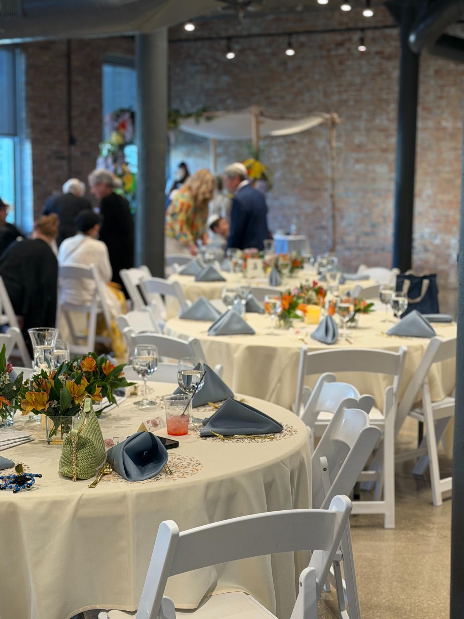 Banquet hall set for a formal reception with round tables, floral centerpieces, and folded napkins; blurred guests in the background.