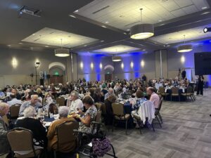 Banquet hall full of guests seated at round tables, dining at a formal event with blue wall lighting.
