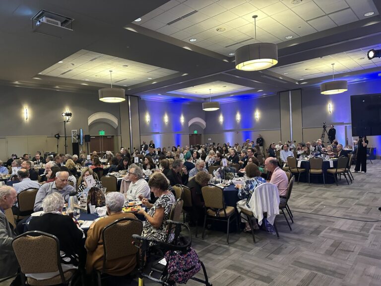 Banquet hall full of guests seated at round tables, dining at a formal event with blue wall lighting.